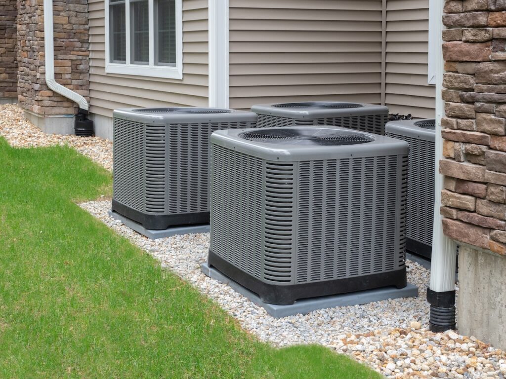 A row of three grey residential central AC condenser units installed on concrete pads and gravel along the side of a house with beige siding and stone accents.