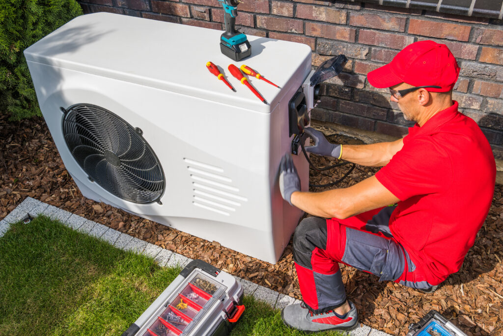 how much to replace a heating and air conditioning system Professional Middle Aged HVAC Technician in Red Uniform Repairing Modern Heat Pump Unit. House Heating and Cooling System Theme.