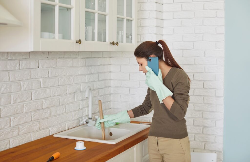 Focused young woman wearing rubber gloves using cup plunger to unclog sink in kitchen while talking on phone. Busy female homeowner calling emergency or plumbing service while solving plumbing issue.