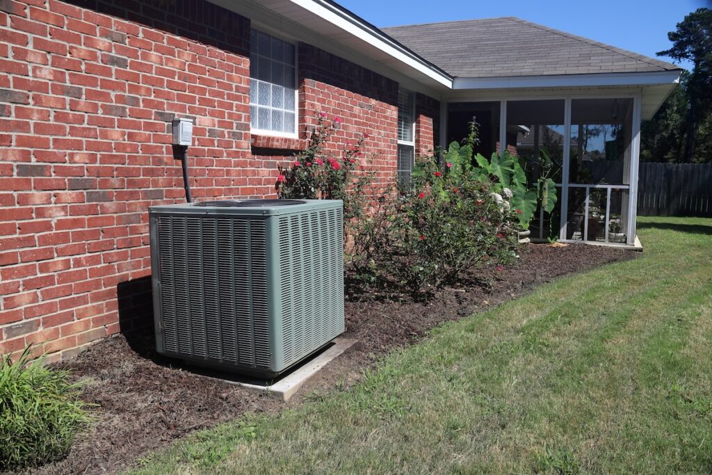 A high-efficiency outdoor condenser unit positioned alongside a classic red brick residential exterior, representing a precision central air installation performed by the licensed HVAC experts at Rolls Mechanical to ensure long-term cooling comfort.