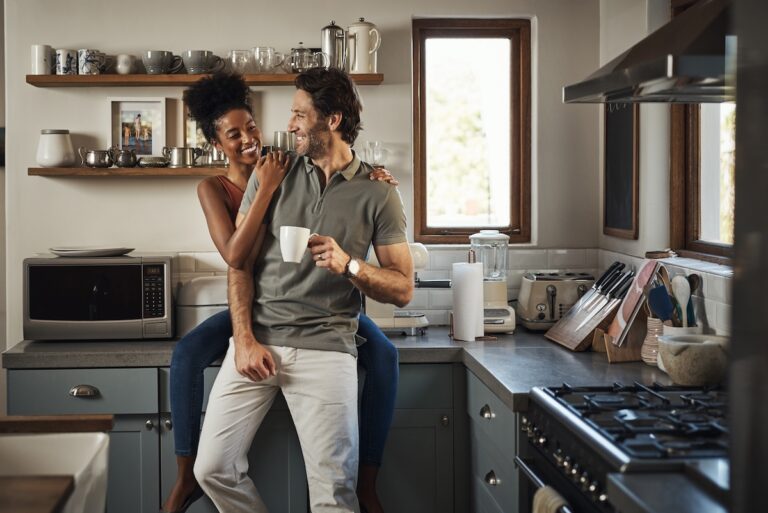 Happy, in love and laughing while an interracial couple enjoys morning coffee and bonding while having good communication in a relationship. Husband and wife talking while standing in kitchen at home.