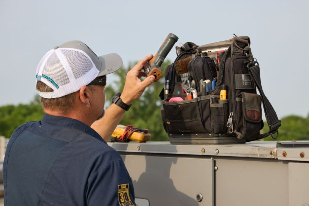 An HVAC professional from Rolls Mechanical selects a diagnostic tool from a fully stocked technician tool bag while performing a routine inspection on a commercial rooftop unit.