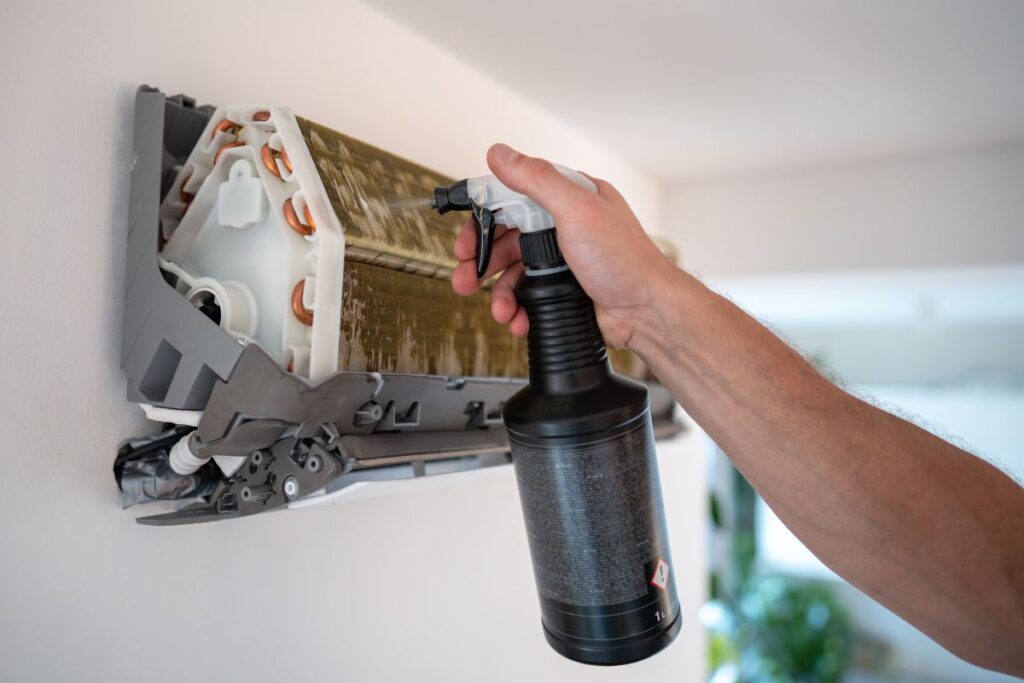 A technician spraying a specialized AC Coil Cleaner foam onto the copper fins of an indoor mini-split unit to remove debris and prevent mold growth.