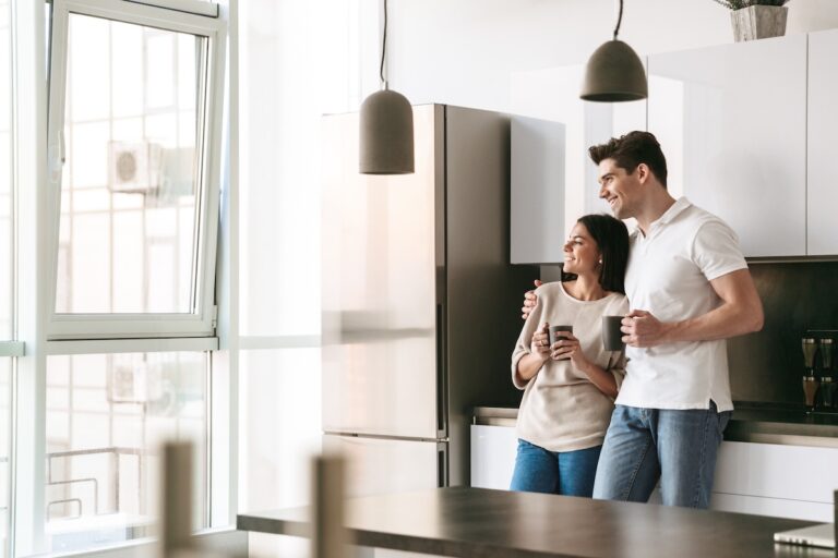 Happy lovely young couple holding cups while standing at the kitchen at home, looking at the window