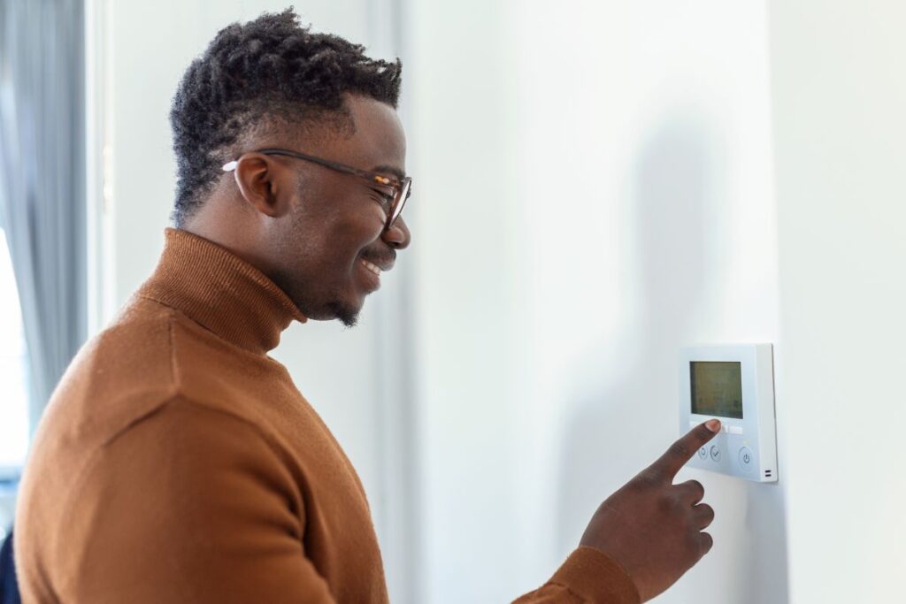 A smiling man adjusting a digital wall thermostat to optimize home heating, an important habit for those wondering how long does a furnace last through consistent temperature regulation.