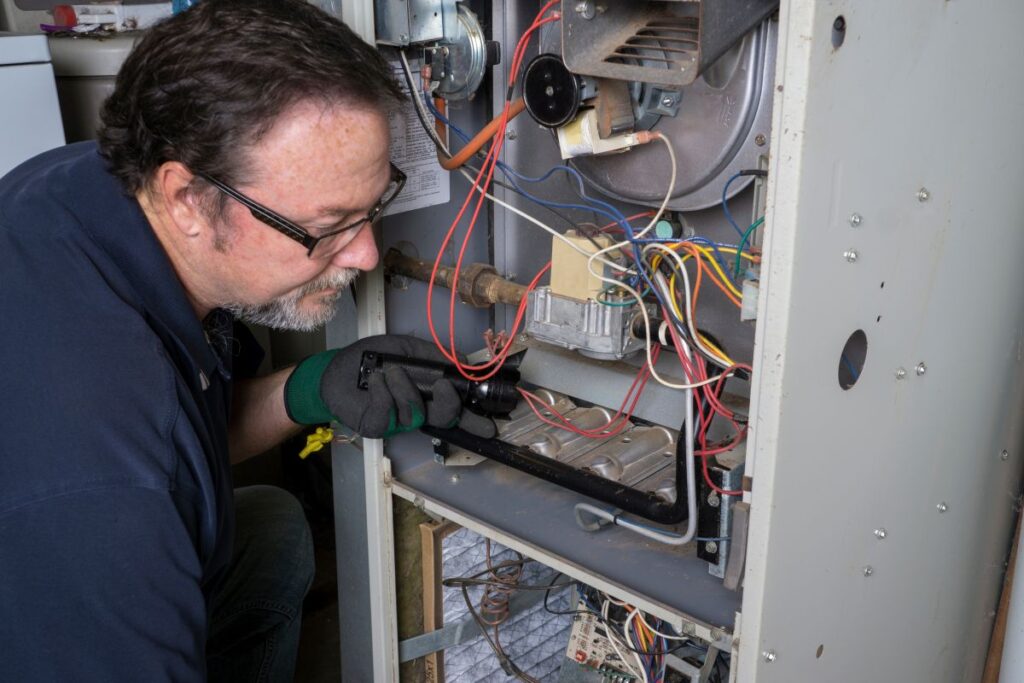 An HVAC technician using a flashlight to inspect the internal burners and wiring of a gas heating system, providing the preventative care that directly impacts how long does a furnace last.