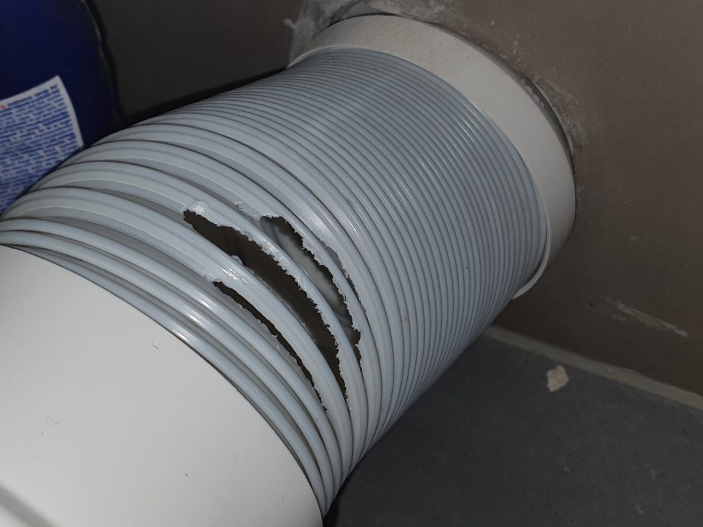A close-up view of a damaged gray flexible corrugated ventilation hose or duct pipe showing a large tear and split in the ribbed plastic material. The flexible tubing appears to be connected to a
